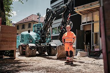 Operatore che indossa indumenti da lavoro ad alta visibilità, in piedi accanto a un escavatore gommato Wacker Neuson EW100 in un cantiere urbano.