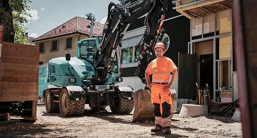 Operator wearing high-visibility workwear standing beside a Wacker Neuson EW100 wheeled excavator at an urban construction site.