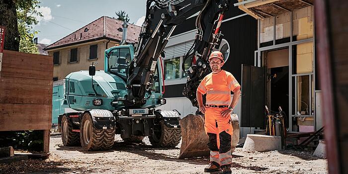 Operatore che indossa indumenti da lavoro ad alta visibilità, in piedi accanto a un escavatore gommato Wacker Neuson EW100 in un cantiere urbano.