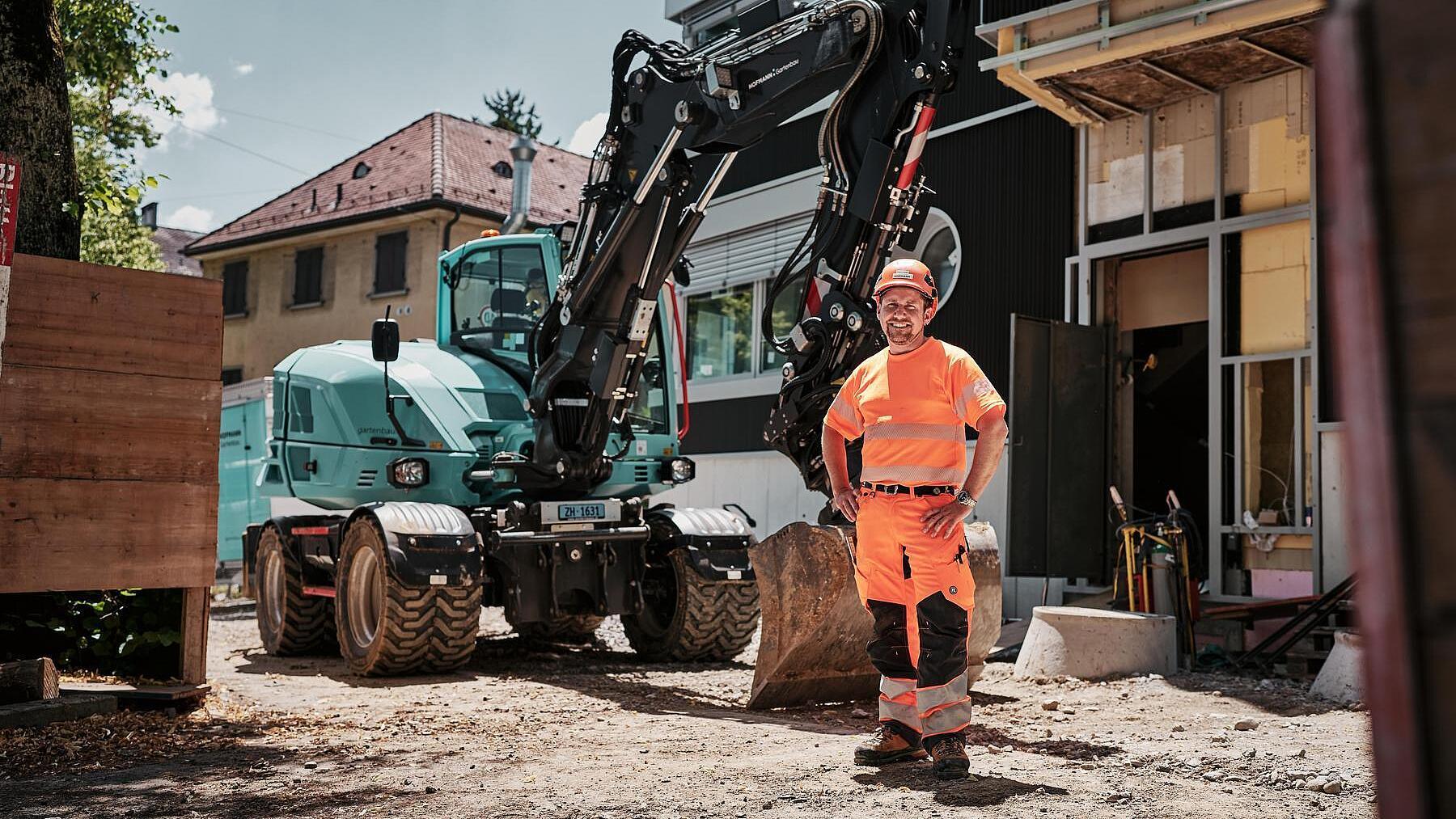 Operatore che indossa indumenti da lavoro ad alta visibilità, in piedi accanto a un escavatore gommato Wacker Neuson EW100 in un cantiere urbano.