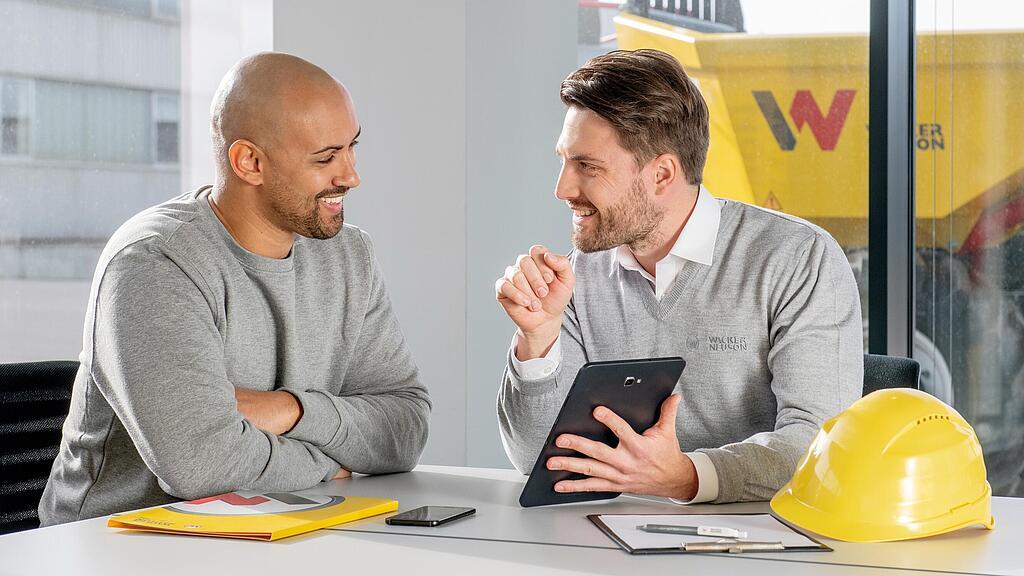 Il dipendente Wacker Neuson sta seduto alla sua scrivania e mostra ai clienti qualcosa sul tablet.