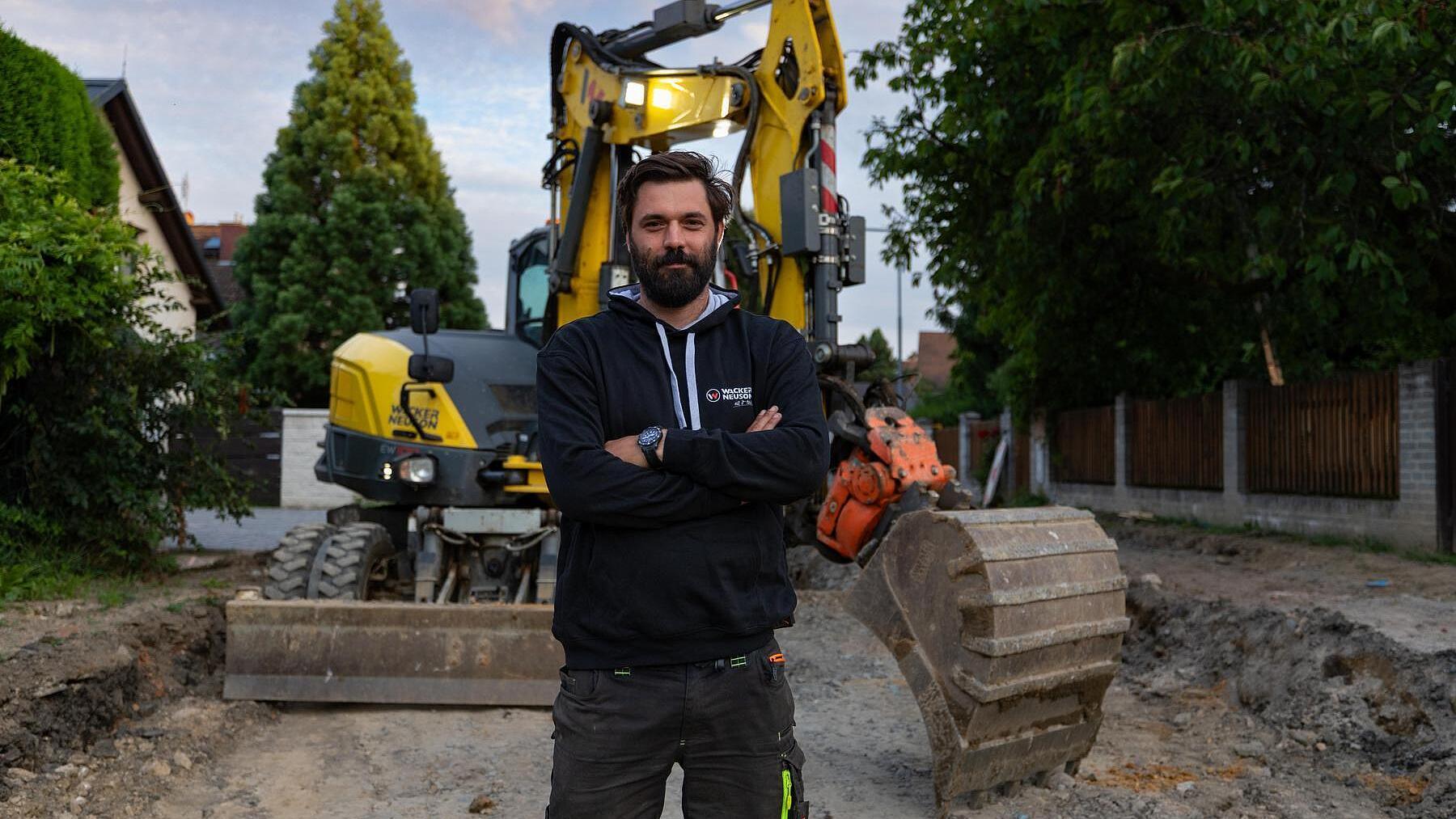 Un professionista del settore edile in piedi in un cantiere davanti a un escavatore gommato Wacker Neuson EW100.